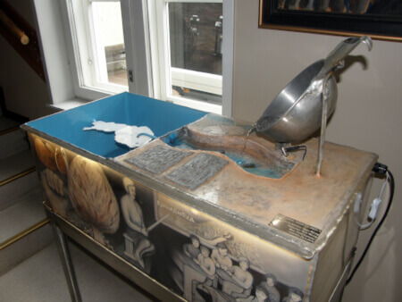 Vesiallas, jonka sivulla maalauksia Kauhanojan koulusta ja yläpuolella vesi valuu kauhasta 'Loimijokea' pitkin Suomen kartaan Kauhanojan kohdalle