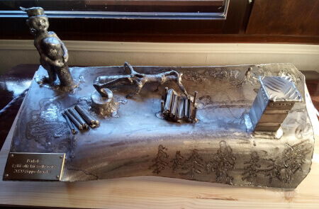 Alumiinista hitsattu taideteos, jossa ukko repii puuta metsässä ja kirves on isketty kantoon puukasojen keskellä